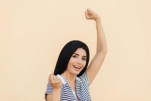 Young happy woman with dark long hair raises fists with joyful expression, has attractive look, wears casual clothes, rejoices her success, has triumph. People, achievement and happiness concept photo