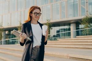 Stylish businesswoman with laptop drinking coffee from disposable paper cup while going at work photo