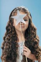 Indoor shot of good looking small female child with long curly hair, wears crown, covers face with magic wand in form of star, isolated over light blue background. Childern and magic concept photo