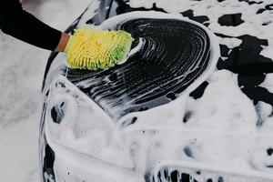 Mans hand with mop cleaning car with auto shampoo. Soap suds on black bonnet. Vehicle concept. Modern automobile covered by foam