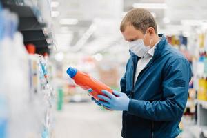 Man makes shopping in supermarket, holds bottle of washing detergent, reads product information, wears protective medical mask and gloves during coronavirus outbreak, chooses something for household photo