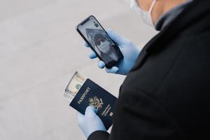 Shot of unrecognizable man follow rules of social distancing, makes video call, holds mobile phone, passport with money wears protective gloves walks outdoor at street. Avoid contact with other people photo