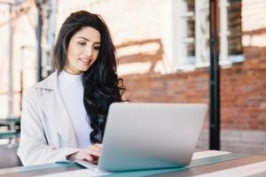 Adorable brunette woman in white clothes sitting outdoors at cafe using her laptop computer communicating with friends online using free internet connection. People, lifestyle, technology concept photo