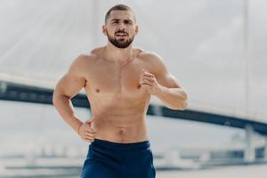 Half length shot of serious male jogger runs actively outdoor concentrated into distance poses with naked torso poses against blurred background with bridge. Fit muscular man runner trains actively photo