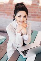 Luxurious brunette female with charming smile having pleasant look into camera while sitting at cafe in front of opened laptop chatting with friends or her boyfriend. People and technology concept photo