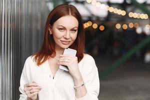 Photo of prosperous female business worker has break after work, sends text messages, holds cell phone in hand, dressed in white shirt, has browh hair, stands outdoor against blurred background