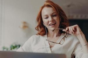 Confident pleased redhead woman concentrated in laptop computer, watches video or movie on laptop computer, enjoys friendly video call, holds transparent glasses, uses free internet connection photo