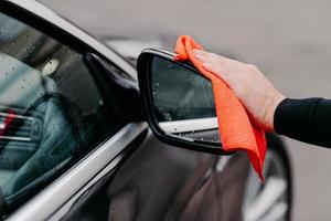 Close up of mans hand wiping water on black car with microfiber cloth. Focus on auto side mirror. Transporation self service