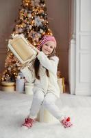 Vertical portrait of delightful small female child in white clothes and warm socks holds big present box, recieves gift from parents on New Year, poses against decorated Christmas tree photo