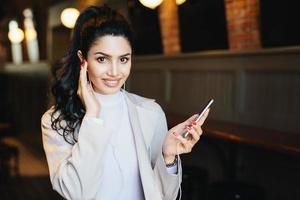 Cute woman with dark pony tail having long eyelashes and expressive eyes sitting at cafe listening to music online enjoying high-speed internet connection. People, beauty and technology concept photo