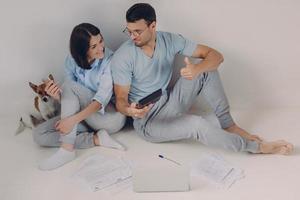 Photo of satisfied man wears spectacles and casual clothes, shows figures on calculator, demonstartes okay gesture, rejoices high profits, works together with female colleague, sit on floor.