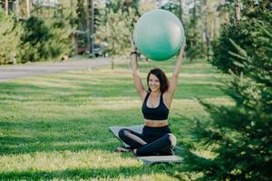 Horizontal shot of brunette fit woman in active wear sits in lotus pose on karemat holds fitness ball over head has gymanstic exercises outdoor during sunny day, smiles pleasantly, enjoys nature
