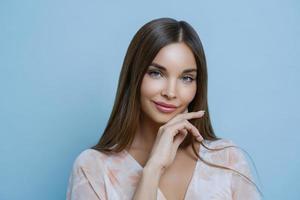 Alluring pleasant looking woman with long dark straight hair touches jawline, looks forward to special event, takes part in profesional photoshoot, has healthy clean skin, poses over blue background photo