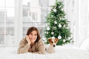 Relaxed beautiful dark haired young woman poses on bed with favourite pet, enjoys bedding time, has lazy day during winter holidays, decorated New Year tree behind, big windows. Cozy atmosphere photo