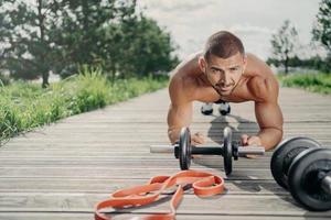 Strong motivated handsome man stands in plank pose, makes abdominal exercises, poses near sport equipment, listens music in wireless earphones, has workout outdoor, looks with serious expression photo