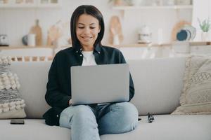 niña feliz escribiendo en la computadora y sonriendo. freelancer está sentado en un cómodo sofá con una computadora portátil. foto