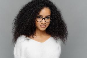 Close up shot of dark skinned female has satisfied expression, bushy curly hair, wears spectacles and white jumper, looks straightly at camera, models over grey background. Natural beauty concept photo