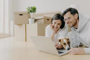 Glad young female and male discuss home repair projects, look attentively into laptop, their dog lies near, pose in living room on floor, dressed in casual clothes. People and new home concept photo