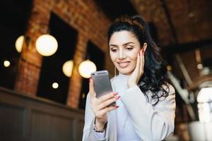 Lovely lady with dark hair, make-up and wonderful manicure wearing white jacket holding mobile phone texting SMS listening to music online on earphones, enjoying free wireless internet connection photo