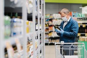 Photo of man in protective medical mask and rubber gloves to prevent coronavirus, poses in supermarket, holds glass bottle of alcoholic drink, reads label, makes shopping during quarantine time