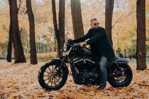 Thoughtful biker rides fast motorcycle, turns away, notices something into distance, wears sunglasses, poses in autumnal park, drives in nature. Lonely motorcyclist poses outdoor at nature during trip
