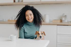 Pretty young lady with Afro hairstyle plays with dog, wears comfortable jumper, drinks coffee or tea in kitchen, going to have walk together, rejoice good weather outdoor. People, animals, spare time photo