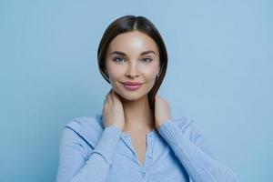 Headshot of young lovely woman keeps hands on neck, has European appearance, happy to hear pleasant words, wears casual sweater, isolated over blue background. Human face expressions concept photo