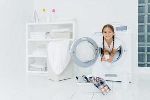 Small girl with appealing appearance, has fun and poses inside of washer, holds detergent, prepares for washing, basin with clothes for putting into washing machine on white floor. Laundry day at home photo