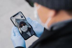 Unrecognizable man has video conversation outdoor, holds mobile phone in protective rubber gloves, wears protective mask because of coronavirus epidemic spread, talks to friend, discuss news photo
