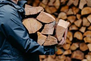 Close up of unrecognizable man dressed in jacket carries bundle of firewood, going to make fire outdoor. Winter is coming. photo