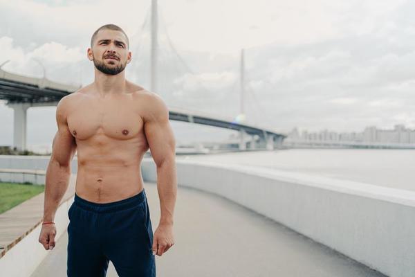 Handsome muscular man with naked torso has outdoor fitness workout looks into distance, has sexy body, dressed in sports trousers stands outside near river bridge image
