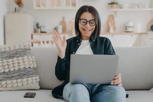 Smiling female waving hand to laptop greeting teacher at lesson, sitting on couch at home. Elearning photo