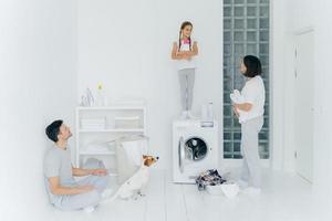 Photo of husband and wife, their dog and daughter do housework in laundry room, little girl stands with detergent on top of washing machine, busy washing clothes and linen, have spring cleaning