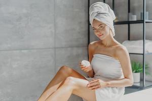 Horizontal shot of pretty woman applies cosmetic cream on skin, poses wrapped in towel, undergoes beauty procedures after taking shower, poses at home in bathroom. Daily hygiene and skin care concept photo