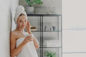 Pleased attractive woman has natural beauty, glowing smooth healthy skin, touches face, enjoys spa procedures, wears white bath towel on head and around body, drinks tea, poses in cozy bathroom photo