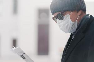 Sideways shot of man in hat, sunglasses and protective mask, reads newspaper outdoor, poses at street, finds out news from press during coronavirus outbreak and quarantine, respiratory virus photo