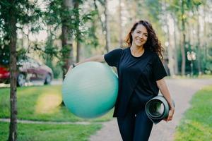 Pleased brunette woman dressed in black t shirt, holds rolled up karemat and fitness ball, prepares for aerobic exercises, smiles pleasantly, poses against nature background. People and sport concept photo