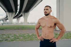 Photo of shirtless handsome bearded man with naked torso poses outdoor keeps hands on waist concentrated into distance goes in for sport, has morning workout, proud to be strong, has perfect abs