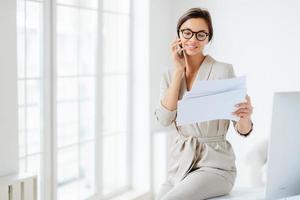 atractiva mujer de negocios lee papeles o documentos comerciales, tiene una conversación telefónica con un socio comercial, examina el papeleo antes de reunirse, se sienta en el escritorio, sonríe alegremente, trabaja con documentación foto