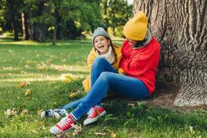 una niña divertida y su madre se sientan juntas cerca de un árbol, caminan en el parque de otoño, disfrutan juntos de un tiempo maravilloso e inolvidable. amable madre y niña pequeña, relajarse al aire libre. concepto de relajación foto