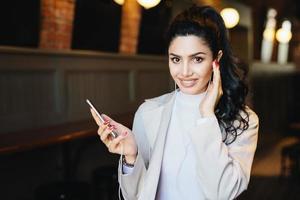 Cute woman with dark pony tail having long eyelashes and expressive eyes sitting at cafe listening to music online enjoying high-speed internet connection. People, beauty and technology concept photo