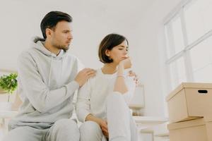 Photo of sad woman complains about problems, man calms, embraces wife, tired after bringing many boxes with things to their new home, wear casual clothes, being stressful. Family problems concept