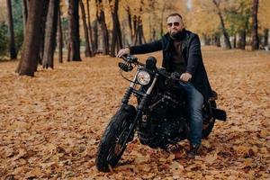 Travel, speed, freedom concept. Fashionable male motorcycle driver poses on black motorbike, wears protective sunglasses, black jacket and shoes, rides in beautiful yellow park during autumn time.