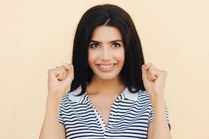 Candid shot of cheerful brunette young female has big hope, keeps hands in fists, believes in better, stands against beige background, has pleasant smile on face. People and happiness concept photo