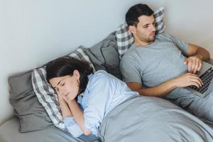 el hombre ocupado se acuesta en la cama, trabaja en una computadora portátil, tiene una expresión seria mientras su esposa tiene un sueño saludable, estando juntos en el dormitorio. dos personas en la cama. gente, familia, concepto de hogar foto