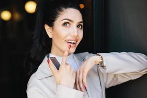 Dreamy beautiful brunette model with curling long eyelashes, dark bright eyes and red manicure dressed in white jacket holding her finger on lips looking up while dreaming about something pleasant photo