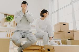 Worried couple pose near stack of cardboard boxes, have to move in other flat, have no money for rent, have dejected sad expressions, sit together in white room, have family conflict and divorce photo