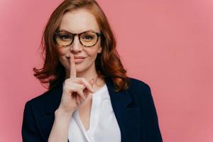 Isolated shot of attractive secret woman keeps fore finger over lips, demonstrates shush gesture, tells secret information, has healthy skin, manicure, makeup, wears formalwear, poses over pink wall photo