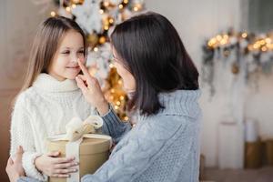 Beautiful female child with long hair, wears white warm sweater, looks in mothers eyes, glad to recieve present on Christmas, celebrate winter holidays in family circle. Happy mother and daughter