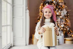 Indoor shot of pleasant looking small kid with blue charming eyes, wears santa hat, holds present in wrapped box, sits over decorated New Year background. Childhood, celebration concept photo
